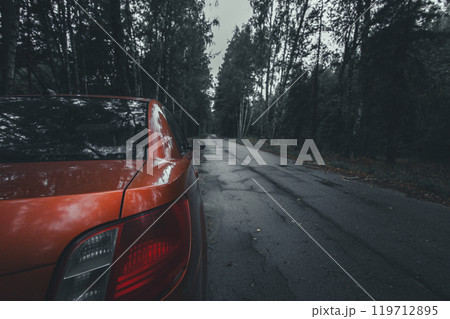 A red car with headlights on, parked on the side of a deserted road leading through a forest on a cloudy day, conveys a sense of mystery and adventure. Car on the side of the road. 119712895