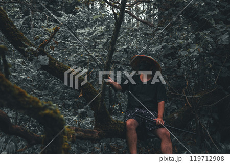 Warrior with katana in the forest. A mysterious man in a black mask and straw hat sits on a fallen tree in the forest, holding a katana, blending into the shadows, creating intrigue. 119712908