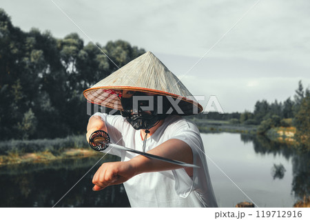 A samurai warrior wearing a conical hat and a black mask unsheathes a katana by a river, demonstrating traditional Japanese martial arts and warrior culture. The warrior practices with a katana. 119712916