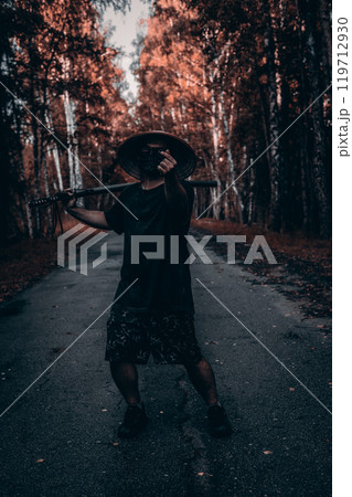 A man in a conical hat and mask stands on a road in the forest, holding a katana on his shoulder and gesturing with his hand. Man with katana in the forest. A man in a conical hat and mask stands on a road in the forest, holding a katana on his shoulder and gesturing with his hand. Man with katana in the forest. 119712930
