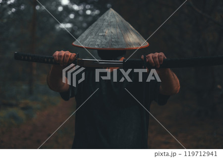 A mysterious samurai with a conical hat and a katana in his hands in the forest. The samurai with the katana is shrouded in shadow, which creates an atmosphere of mystery and intrigue. 119712941