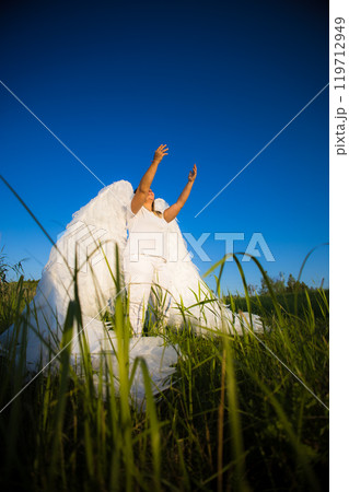 A woman in white clothing and with large white feathers on her wings raises her hands to the sky while standing in a grassy field on a sunny day. The angel asks the sky for help. 119712949