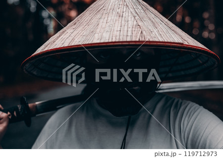 A mysterious man in a conical hat and mask confidently holds a katana in a serene forest, radiating strength and danger. A fearless samurai warrior stands with a katana on his shoulder. A mysterious man in a conical hat and mask confidently holds a katana in a serene forest, radiating strength and danger. A fearless samurai warrior stands with a katana on his shoulder. 119712973