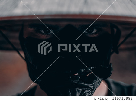 A fierce look of a samurai in a mask and hat. In a dimly lit forest, a man wearing a black mask and a traditional Asian conical hat looks at the camera with a masculine and strong expression. 119712983