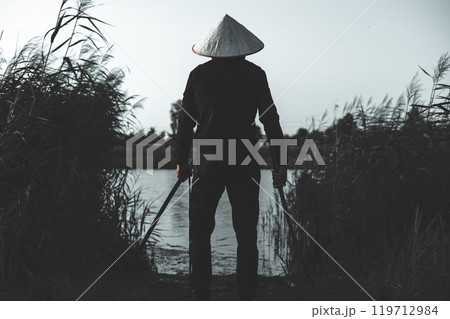 A samurai warrior wearing a conical hat stands on the bank of a river at dusk, holding a katana, creating a dramatic and mysterious scene. A samurai stands on the bank of a river with a katana. 119712984