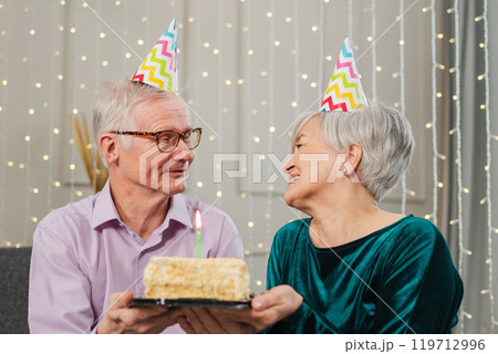 Make a wish. Family senior couple in party hat cap celebrating birthday anniversary together at home. Old woman blowing out burning candles on birthday cake. Old man wishes wife happy birthday Make a wish. Family senior couple in party hat cap celebrating birthday anniversary together at home. Old woman blowing out burning candles on birthday cake. Old man wishes wife happy birthday 119712996