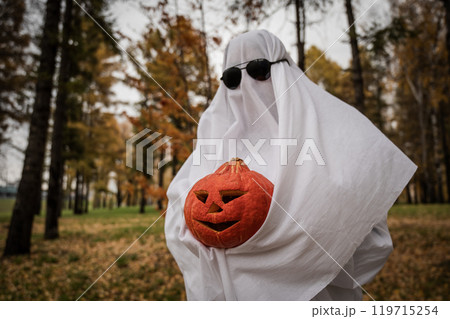 Woman in white sheet holding jack o'lantern against autumn forest background. Halloween costume. Woman in white sheet holding jack o'lantern against autumn forest background. Halloween costume. 119715254