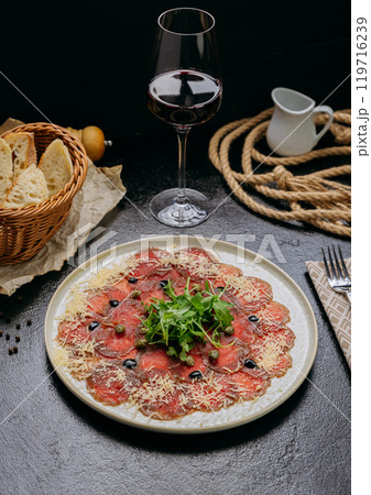 Beef carpaccio with arugula, Parmesan, capers, and olives, served with red wine and fresh bread 119716239
