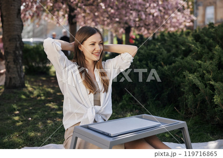 Cherry blossoms, Blossoms woman, Park cherry. Seated in park beneath cherry blossoms, woman leans back with a smile, hands resting behind her head as she uses her laptop. Cherry blossoms, Blossoms woman, Park cherry. Seated in park beneath cherry blossoms, woman leans back with a smile, hands resting behind her head as she uses her laptop. 119716865