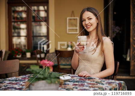 holding coffee cup, Outdoor Coffee, Female Smiling. Young woman smiles while holding coffee cup at outdoor cafe, creating warm and relaxed atmosphere. 119716893