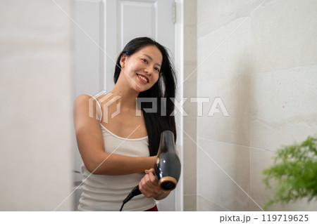 A smiling Asian woman is looking in the mirror, using a hair dryer in the bathroom after shower. A smiling Asian woman is looking in the mirror, using a hair dryer in the bathroom after shower. 119719625