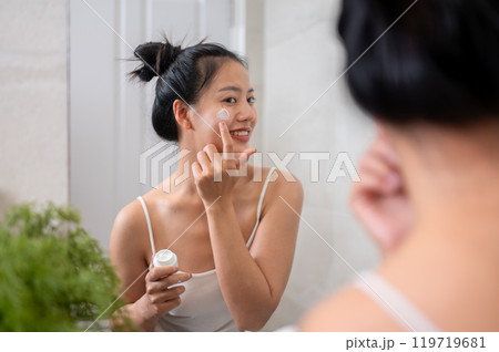 A natural Asian woman is applying facial cream in front of the mirror in the bathroom after shower. 119719681