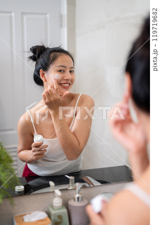 A natural Asian woman is applying facial cream in front of the mirror in the bathroom after shower. A natural Asian woman is applying facial cream in front of the mirror in the bathroom after shower. 119719682