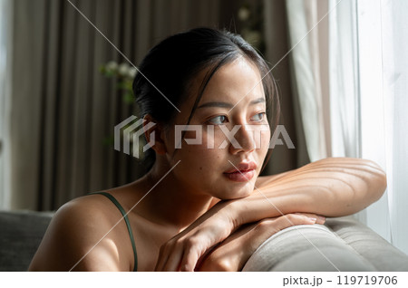 A beautiful, thoughtful woman is sitting on a sofa at home, looking out the window, deep in thought. A beautiful, thoughtful woman is sitting on a sofa at home, looking out the window, deep in thought. 119719706