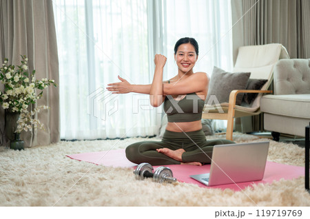 A sporty Asian woman in sportswear is stretching her body on a yoga mat, exercising at home. 119719769
