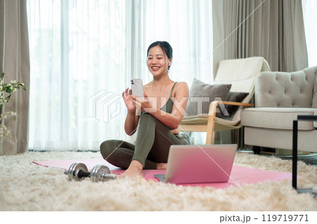 A beautiful, positive Asian woman in sportswear is resting on a yoga mat after her workout at home. 119719771
