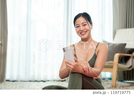 A beautiful, positive Asian woman in sportswear is resting on a yoga mat after her workout at home. 119719775