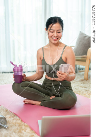 A beautiful, positive Asian woman in sportswear is resting on a yoga mat after her workout at home. 119719776