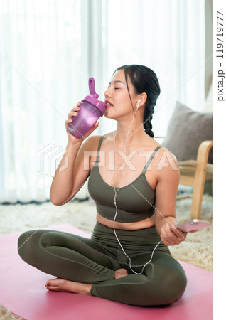 A sporty Asian woman in yoga clothing is sipping water from a bottle after her yoga session at home. A sporty Asian woman in yoga clothing is sipping water from a bottle after her yoga session at home. 119719777