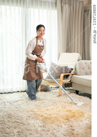 A beautiful Asian woman is cleaning a carpet with a vacuum cleaner in the living room at home. 119719809