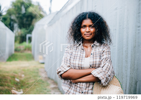 Portrait of a confident woman farmer in a checkered shirt and apron arms crossed holding a hat smiling in front of a farm's lush greenhouse tomatoes. Nature's beauty in a farming setting. Portrait of a confident woman farmer in a checkered shirt and apron arms crossed holding a hat smiling in front of a farm's lush greenhouse tomatoes. Nature's beauty in a farming setting. 119719969