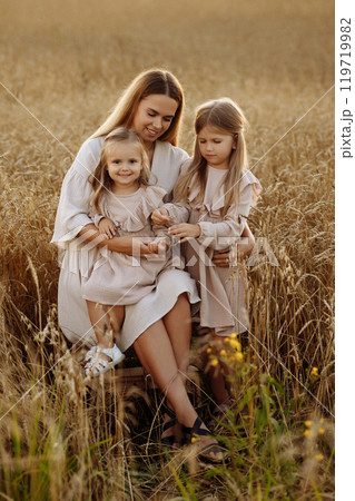 Young woman with two daughters in a wheat field during sunset. 119719982