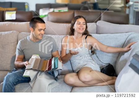 In store wife sit with legs on gray sofa to test in use,husband examines upholstery fabric In store wife sit with legs on gray sofa to test in use,husband examines upholstery fabric 119721592