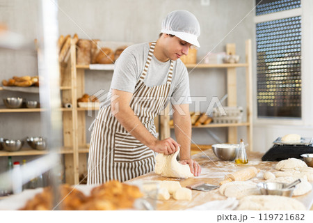 Guy baker in uniform prepares raw dough, kneads it with hands 119721682