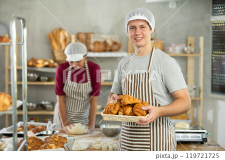 Portrait of young happy male baker working in bakehouse, holding tray with fresh bakery goods on kitchen 119721725