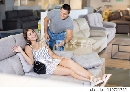 Couple chooses furniture in store.Wife is sitting on sofa,husband is standing next to her Couple chooses furniture in store.Wife is sitting on sofa,husband is standing next to her 119721735