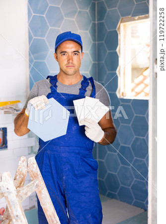 Skilled builder in work clothes posing with ceramic tiles and construction tools while repair and decoration in apartment 119722258