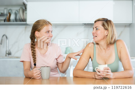 Cheerful mother and teenage daughter drinking tea and talking in kitchen 119722616