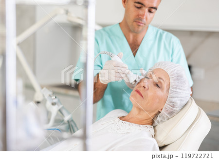 Cosmetologist performing anti-aging face hardware therapy procedure for aged female client in clinic of aesthetic medicine Cosmetologist performing anti-aging face hardware therapy procedure for aged female client in clinic of aesthetic medicine 119722721