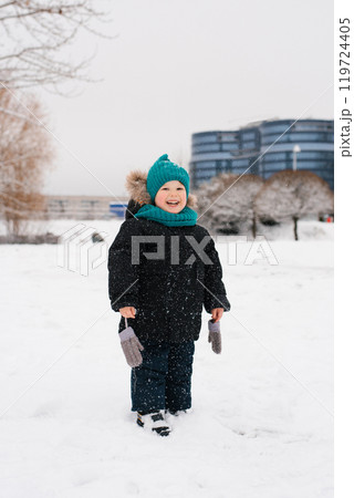 Boy child is standing in the snow wearing a black coat and a blue hat. The child is smiling and holding two mittens. The scene is set in a snowy park, with a building in the background Boy child is standing in the snow wearing a black coat and a blue hat. The child is smiling and holding two mittens. The scene is set in a snowy park, with a building in the background 119724405