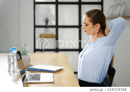 Woman stretching at a desk with a laptop during a work break in a modern office setting Woman stretching at a desk with a laptop during a work break in a modern office setting 119724598