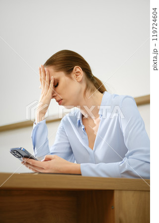A woman sitting at a desk appears stressed while looking at her smartphone in a light-filled room 119724604