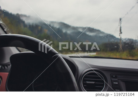 Driving in mountains on rainy day. Car dashboard with steering wheel with rain drops on glass and autumn mountains on background. Car on highway in mountains. Autumn vacations concept. Driving in mountains on rainy day. Car dashboard with steering wheel with rain drops on glass and autumn mountains on background. Car on highway in mountains. Autumn vacations concept. 119725104