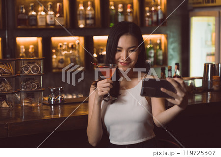 Asian woman taking a photo of herself while drinking whiskey at the bar. 119725307