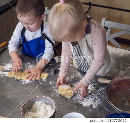 Two young kids in aprons make cookies on a flour-covered kitchen counter, surrounded by cookie cutters and bowls. Laughing and kneading dough, they fill the kitchen with the joy of homemade cookies. 119728081