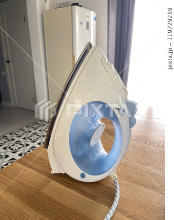 White and blue iron stands upright on a wooden table in a modern kitchen setting, capturing the essence of domestic chores. Natural light highlights the sleek design and functionality of the everyday 119729289