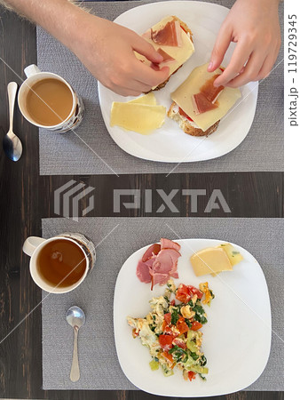 Cozy breakfast setting shows a person preparing open faced sandwiches while another plate displays a colorful scramble with coffee cups nearby. Portrays a homey and inviting morning meal Cozy breakfast setting shows a person preparing open faced sandwiches while another plate displays a colorful scramble with coffee cups nearby. Portrays a homey and inviting morning meal 119729345
