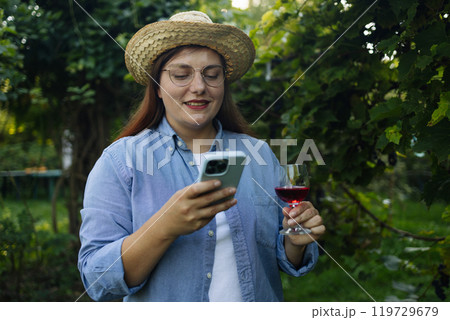 Portrait view of Caucasian woman in a straw hat with a phone is tasting red wine in a vineyard. Beautiful young woman with a glass of wine. Happy winemaker drinking wine after successful grape harvest 119729679