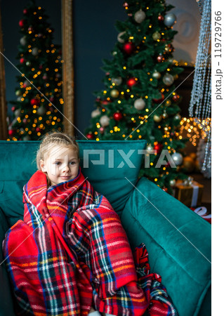 Little girl sitting wrapped in a warm red blanket against the background of a Christmas tree with red golden toys and a glowing garland at home 119729766