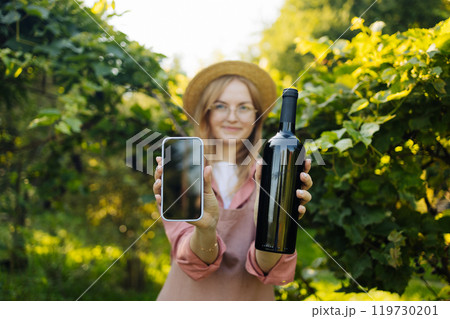 Caucasian woman vintner Shows smartphone and bottle of red wine to camera enjoying wine in her vineyard. Female in hat and an apron holding red wine bottle outdoors. Winery at autumn 119730201