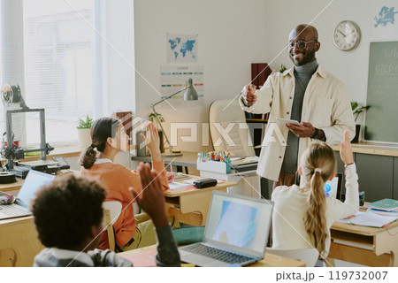 Educator explaining a concept in bright classroom while students attentively listen and raise hands. Classroom environment includes books, laptops, and educational posters Educator explaining a concept in bright classroom while students attentively listen and raise hands. Classroom environment includes books, laptops, and educational posters 119732007