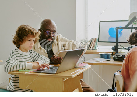 African American teacher mentoring focused young student using laptop in bright classroom. Books and technology visible in background creating an engaging learning environment 119732016