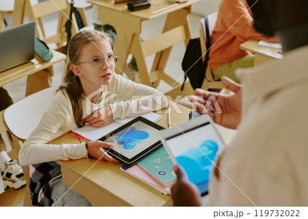 Young girl sitting at desk in classroom holding tablet and engaging with teacher School supplies and other students in background busy with activities 119732022