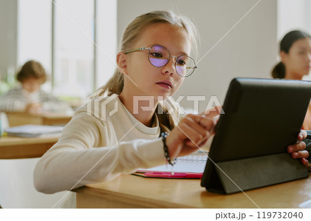 Young girl wearing glasses sitting at wooden desk using tablet in classroom surrounded by fellow students in bright learning environment Young girl wearing glasses sitting at wooden desk using tablet in classroom surrounded by fellow students in bright learning environment 119732040