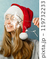 Heart shaped bengal fire in hand of beautiful girl with long red hair in New Year hat on grey background of house.  119732233