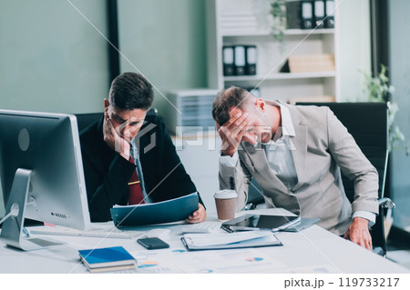 Two Unhappy tired businessman, corporate manager, sits at the workplace, tired from online work, stressed, nervous, massages the bridge of his nose, closed his eyes, experiencing a headache. Two Unhappy tired businessman, corporate manager, sits at the workplace, tired from online work, stressed, nervous, massages the bridge of his nose, closed his eyes, experiencing a headache. 119733217